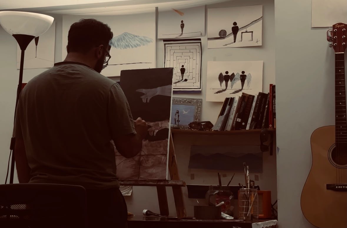 The artist painting at an easel in a studio lined with artworks and books, with a guitar beside the desk.