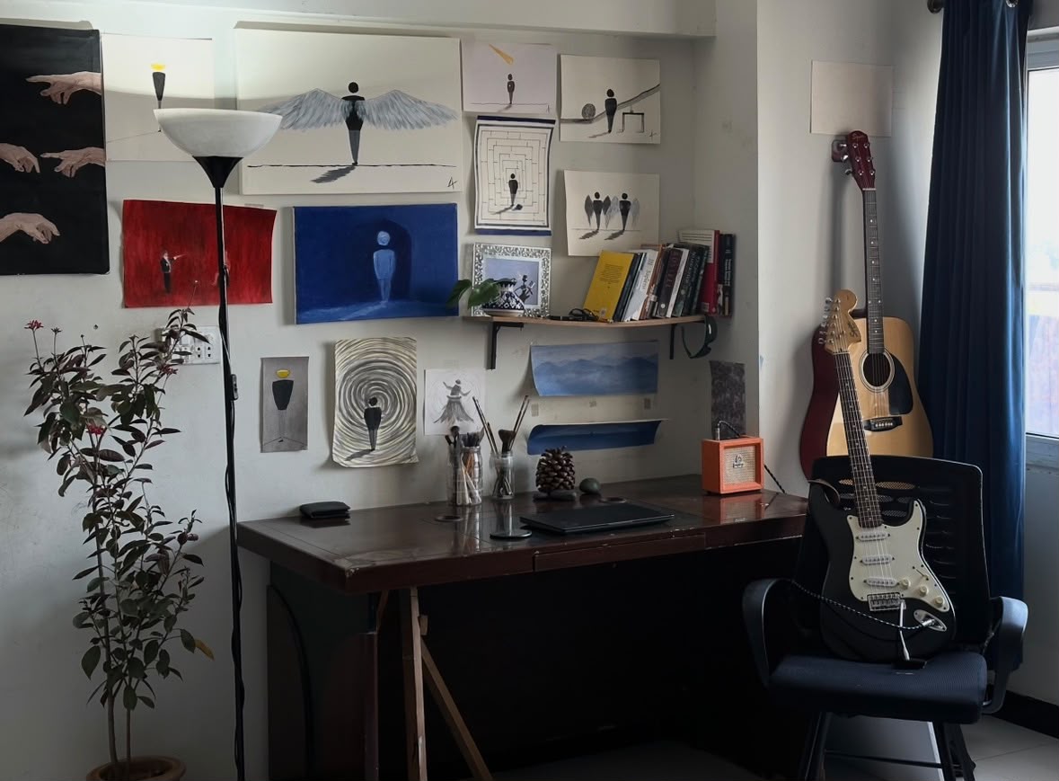 A sunlit studio corner with paintings above a wooden desk, brushes, books, a plant, and two guitars.
